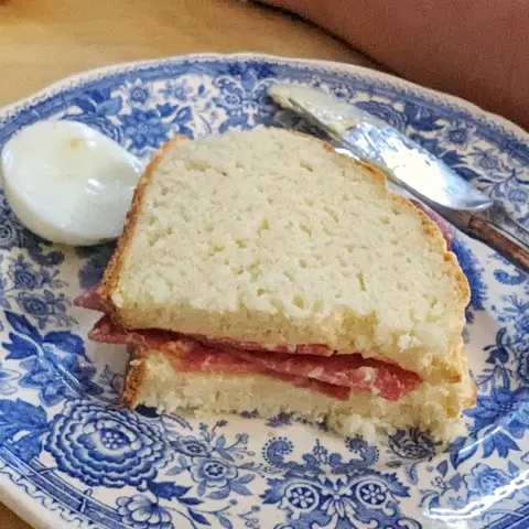 Heller Esstisch, im Fokus ein weißer Teller mit blauem, floralem Motiv (Serie "Burgenland" von Villeroy & Boch). Auf dem Teller liegt ein benutztes Buttermesser, eine Eierkuppe und eine zusammengeklappte Stulle Weißbrot. Die Schnittkante ist sichbar, Salamischeiben sind erkennbar.