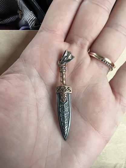 A photo of a 1.5βish sized necklace charm shaped like a sword or dagger. The hilt is a copper color and the blade is grey silver with squared spirals carved into it. I am holding the charm in the palm of my hand.