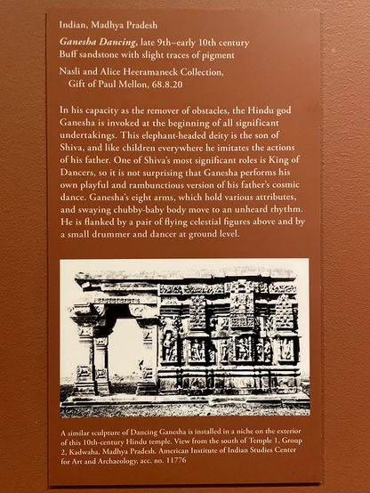 sign: “In his capacity as the remover of obstacles, the Hindu god Ganesha is invoked at the beginning of all significant undertakings. This elephant-headed deity is the son of Shiva, and like children everywhere he imitates the actions of his father. One of Shiva's most significant roles is King of Dancers, so it is not surprising that Ganesha performs his own playful and rambunctious version of his father's cosmic dance. Ganesha's eight arms, which hold various attributes, and swaying chubby-baby body move to an unheard rhythm. He is flanked by a pair of flying celestial figures above and by a small drummer and dancer at ground level. [photo] A similar sculpture of Dancing Ganesha is installed in a niche on the exterior of this 10ch-century Hindu temple. View from the south of Temple 1, Group 2, Kadwaha, Madhya Pradesh. American Institute of Indian Studies Center for Art and Archaeology, acc. no. 11776”