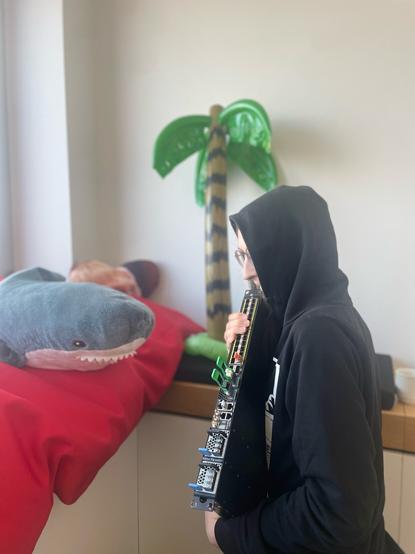A network engineer with a black hoodie, very carefully and lovingly holding a cute 1 RU router. In the background are a Blahaj, an inflatable palm tree and a bean bag.