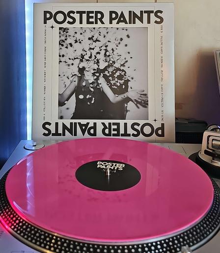 A pink vinyl record sits on a turntable. Behind the turntable, a vinyl album outer sleeve is displayed. The front cover shows a woman and a man with confetti falling from above them, covering their faces