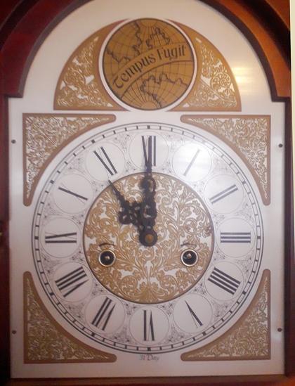 This is the face, or dial, of a Grandfather clock. It is not so old, but is made to look old. 
The whole clock is a long case, made of wood, about my height, and the front of that case is glazed so that you can see the pendulum swaying back and forth inside of it. 
Here we only show the dial, which is a square with the circle of the clock face inside so that the rim touches the middle of each side. This leaves a space in each corner which is filled in with a decorative swirly pattern,  which is repeated in the very middle of the circle, where there are the hands and the winding holes below. Between the border and this decorative circle is a white roundel in which are the clock times in those odd numbers involving entirely the letter I and the letter X. The hands of the clock are set to eleven o`Clock. 
Above the top border of the square is an arch shape with another circle inside it, or rather it is a circle with two decorative sides forming it into an arch, That circle is solid gold (or solid gold paint anyway) and it is a kind of globe design but showing continents I do not know. Across the globe in a banner curving from bottom left to middle right it says Tempus Fugit - which is Latin for Time Flies, rather gloomily.