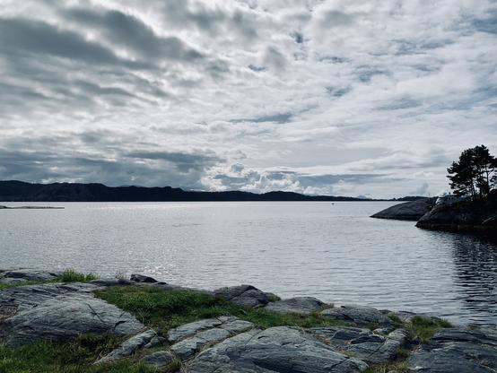 View of an archipelago landscape on the Norwegian west coast. The sky is mostly cloudy, only through a few cloud holes the sun tries to come through. The water surface is smooth and calm. On the right edge, on a peninsula jutting into the picture, is a mobile home.