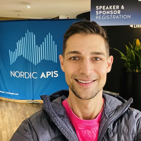 Selfie of Jeremiah smiling wearing hot pink sweater in front of Nordic APIs Speaker registration sign in hotel lobby
