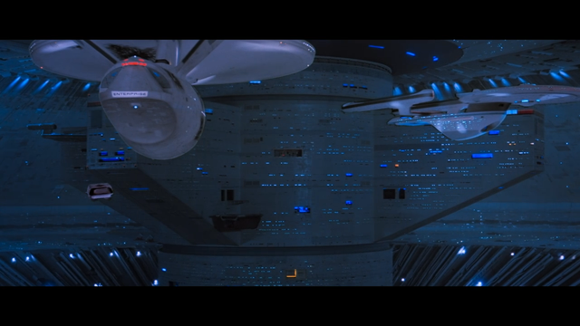 The USS Enterprise prepares to park near the USS Excelsior in the Spacedock. In the background on the left, a study model of a triangular ship made in the mid-70s for the unproduced Star Trek: Planet of the Titans movie project. This design would inspire the USS Discovery decades later.