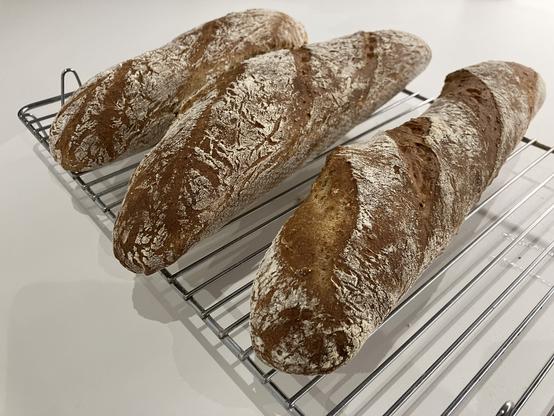 Three golden brown baguettes dry on a metal rack over a white kitchen countertop.