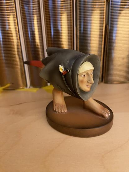 A figurine based on a character from a Hieronymus Bosch painting. It gas a human head in a cloak, a bird tail, and feet, but no legs, arms, or abdomen. In the background, books with gold horizontal stripes reflecting golden light.