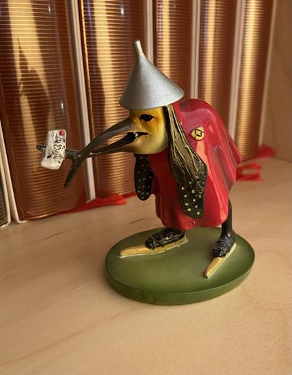 Another figurine based on a character from a Hieronymus Bosch painting. It has a hat like a silver funnel, a long crisscrossed beak with a letter stuck to it, long droopy ears, a red cloak, and ice skates.
