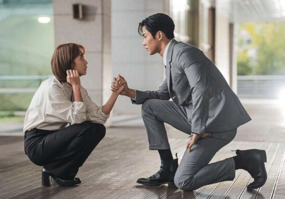 Jo Bo Ah sitting on her heels, holding hands with a kneeling Rowoon dressed in office attire in the 2023 South Korean drama “Destined with You"