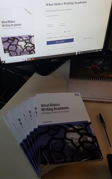 Pile of 'What Makes Writing Academic: Rethinking Theory for Practice' paperbacks by Julia Molinari on a desk with a computer screen as backdrop displaying Bloomsbury's web page of the book.
