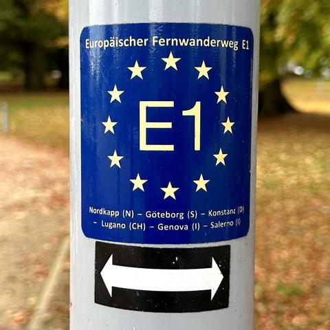Wegzeichen des Europäischen Fernwanderweges E1. Gelbe Schrift „E1“ umgeben von kreisförmig angeordneten gelben Sternen.