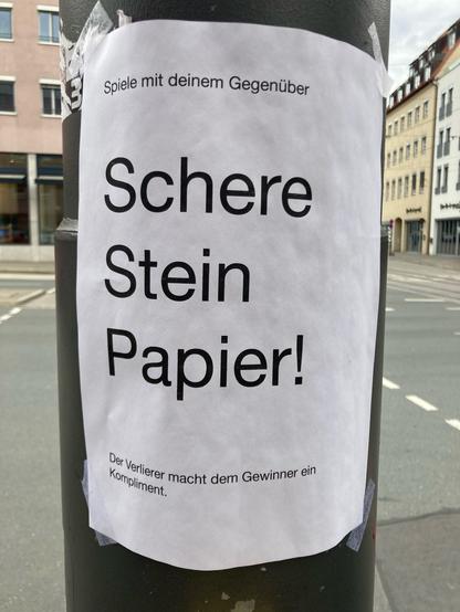 Ein Schild an einer Ampel in Augsburg (Kreuzung Karlstraße / Karolinenstraße). 

Es handelt sich um eine Spielanleitung: 
„Spiele mit deinem Gegenüber Schere, Stein, Papier!
Der Verlierer macht dem Gewinner ein Kompliment.“