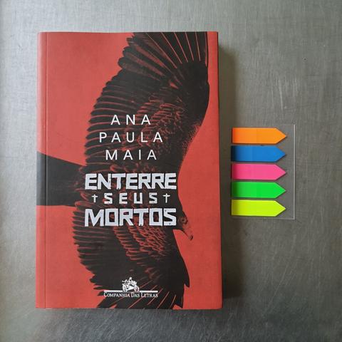 Foto colorida, mostra visto de cima de um balcão de alumínio o livro Enterre Seus Mortos, de Ana Paula Maia, publicado pela editora Companhia das Letras. Ao lado dele uma cartela de post-its coloridos de marcação. Na capa do livro há uma imagem em duotone de uma ave semelhante a um corvo com as asas abertas em pleno vôo.
