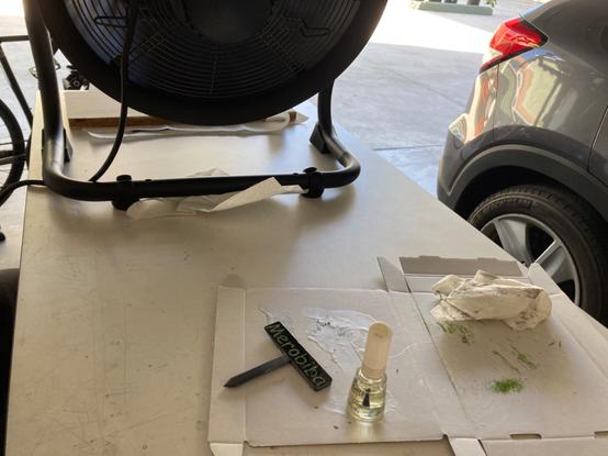 The plant sign, nail polish, and cardboard behind a large fan on the end of my workbench.