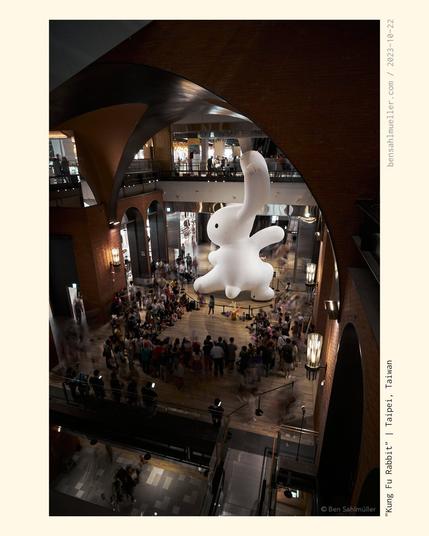 A giant (maybe 15m tall!) white rabbit in the open space of a mall. There is a performance artist standing in front of it, drawing a crowd of kids and families. The Rabbit has one arm raised as if preparing for a kung fu kick.