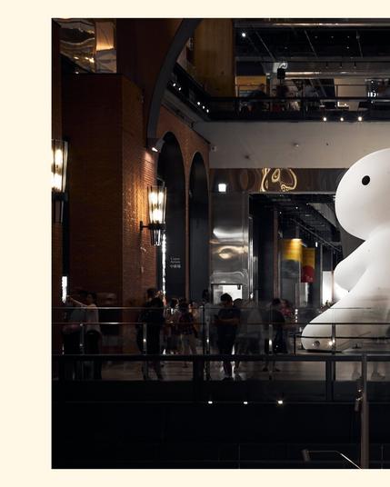 A giant (maybe 15m tall!) white rabbit in the open space of a mall. There is a performance artist standing in front of it, drawing a crowd of kids and families. The Rabbit has one arm raised as if preparing for a kung fu kick.