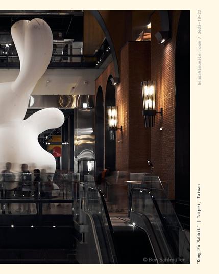 A giant (maybe 15m tall!) white rabbit in the open space of a mall. There is a performance artist standing in front of it, drawing a crowd of kids and families. The Rabbit has one arm raised as if preparing for a kung fu kick.