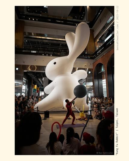 A giant (maybe 15m tall!) white rabbit in the open space of a mall. There is a performance artist standing in front of it, drawing a crowd of kids and families. The Rabbit has one arm raised as if preparing for a kung fu kick.