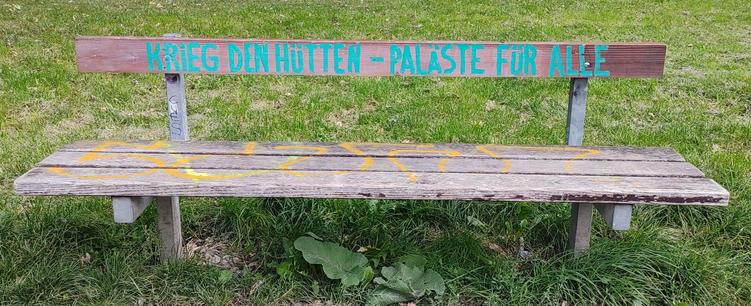 Eine Holzbank vor einer Wiese trägt die grüne Aufschrift "Krieg den Hütten - Paläste für alle"