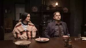 dark room, table and plates. woman with dark-hair and native-american outer clothes looking right towards a man in dark brown clothes looking up and thoughtful