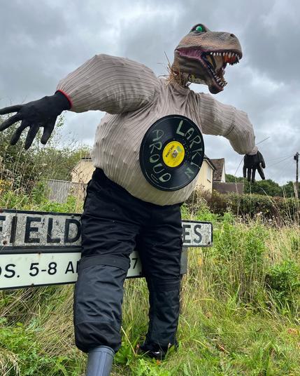 Photograph of a scarecrow standing on field-to-verge margins in rural Buckinghamshire, England, with a Tyrannosaurus Rex mask for a head. It looks terrifying.