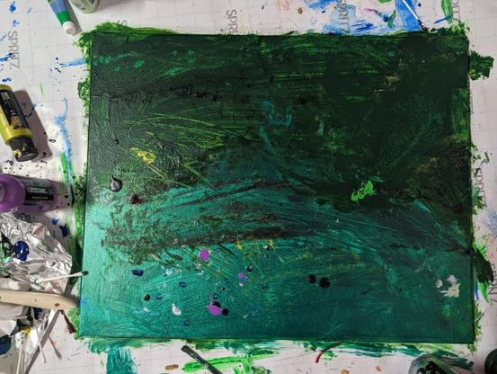 A canvas dries on wrapping paper surrounded by bottles of acrylic paint. The painting is a tumult of various shades of green that are metallic at the bottom and gradient into British racing green at the top. There are spots of other colors that were applied with q-tips but the whole piece is violently green. I love it