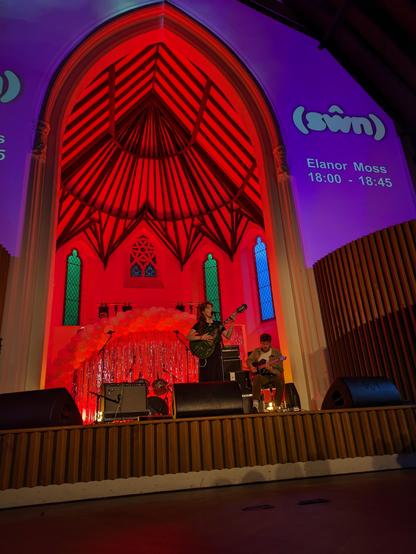 A picture of a singer playing guitar in a church archway with Swn logos projected on the walls nearby.