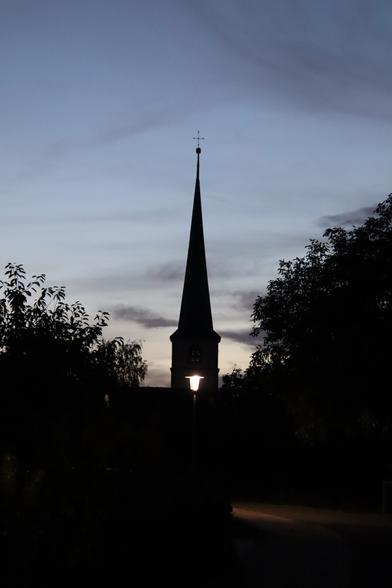 Nachts, Aufnahme eines Kirchturmes links und rechts daneben die Silhouette von Bäumen. Der Himmel ist grau-blau. Eine Strassenlatetne ist mittig im Kirchturm zu erkennen.
Matze-work