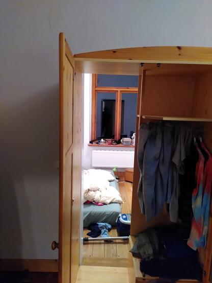 Blick durch einen offenen Schrank in dem an einer Kleiderstange rechts Kleider hängen, links ist die Rückwand entfernt und man blickt in ein kleines Zimmer auf eine auf dem Boden liegende Matratze.