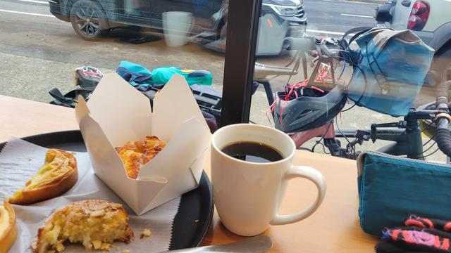 Photo looking out through the window of a pastry shop. On the counter in front of the window, several pastries on a tray, two of them half eaten, and a cup of coffee. Outside the window, two bikes parked together with a bike helmet hanging from the handlebars of one that has a blue bag on its handlebars.