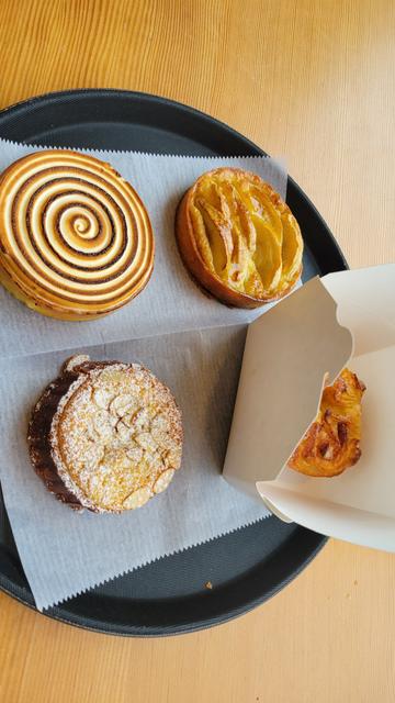 Photo of four pastries on a tray. One has a beautiful white and brown swirled top. Another is dusted with powdered sugar. A third has slices of Apple neatly arranged. The 4th is half hidden inside of white take out box.