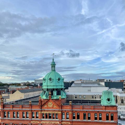 Corner-sited red-brick symmetrical four-storey department store, built 1902 to 1905, on the site of another department store built 1859, with ornate green copper dome and weather-vane. The city recedes in the background. The sky is blue hued above and shot through with skeins and puffs of cloud.