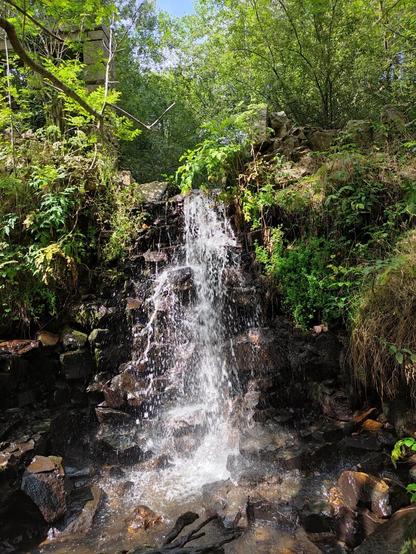 A small waterfall in the middle of the mountains. It has a lot of vegetation around it.