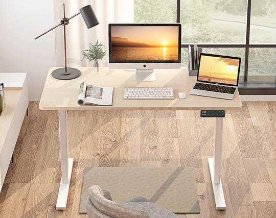 Ein weißer Maidesite T2 Pro Plus-Schreibtisch mit Tischplatte in Eiche steht als Mac-Arbeitsplatz in einem Raum vor einem großen Fenster. Foto (c) Maidesite.