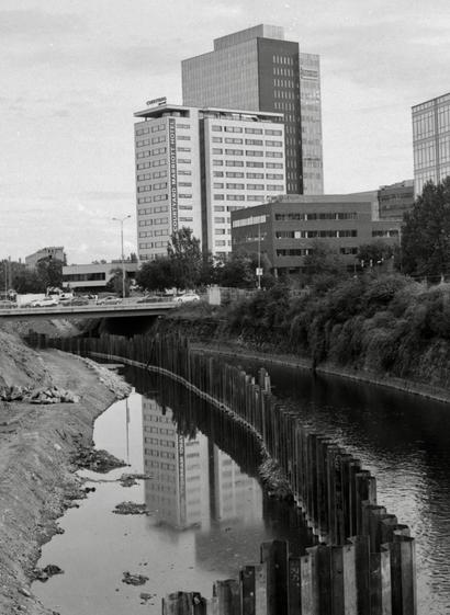 Black and white film photo. River divided by iron plates due to building an embankment. The hotel building in the background is reflected on the surface of the water.