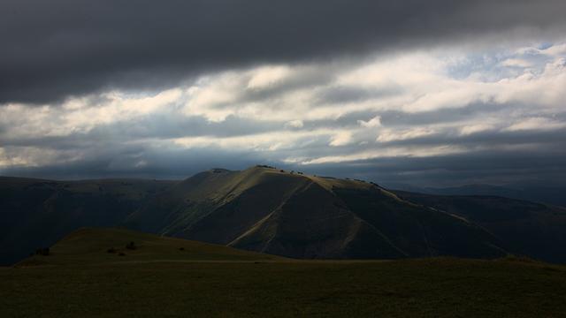View from a meadow in the mountains towards another mountain ridge. The sky is mostly grey but the sun can pass in several locations and so lights up parts of the opposite ridge.