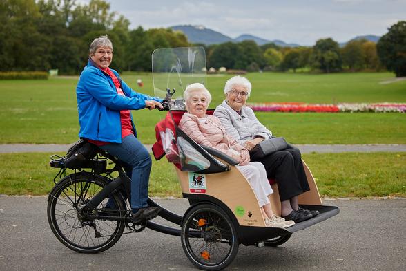 In einer Fahrradrikscha sitzen vorne zwei ältere Damen. Die Rikscha wird gefahren von einer Frau in einer blauen Jacke gefahren. Alle drei Personen lachen. Im Hintergrund sieht man eine Wiese (den Hofgarten in Bonn) und das Siebengebirge.