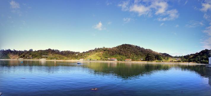 Photo shows a sea bay shaped like a horseshoe with deep blue water and lighter blue sky. It is a sunny day with some light clouds in the sky. 
The landscape in between are small hills and forest with small houses, huts and boat shacks here and there.