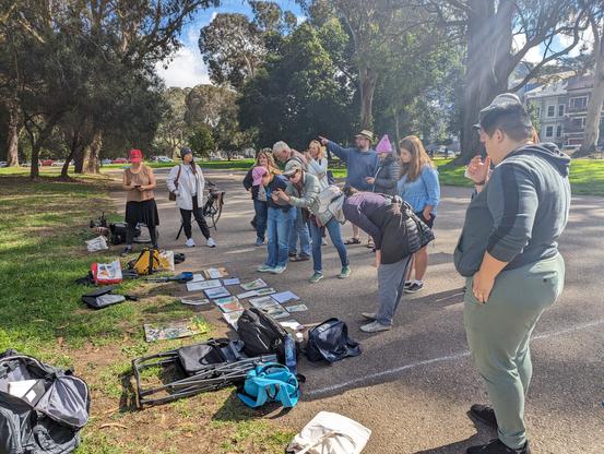 15 artists from the Sunset Sketchers urban sketching group gather around sketchbooks lying on the grass in San Francisco's Panhandle Park to share their work after a meetup.