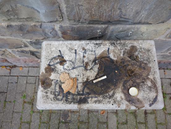 Krume und Rinde einer Scheibe Paderborner Roggenmischbrot sowie ein Zigarettenstummel und ein Kronkorken liegen auf einem Stromkasten in einer Lache einer bräunlichen Flüssigkeit.