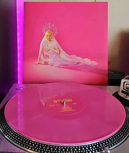 A pink vinyl record sits on a turntable. Behind the turntable, a vinyl album outer sleeve is displayed. The front cover shows Tessa Violet on the floor in an all pink room. She is wearing a see through gown, and a large crown. She is covering her breasts with her left hand and arm.