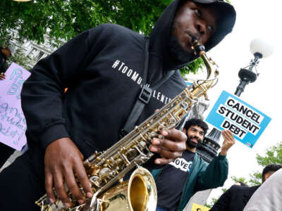 Protest where someone holds a sign the says "Cancel Student Debt" and another person plays a saxophone.