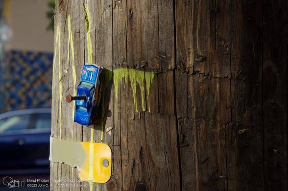 A closeup of a weathered blue toy Hot Wheels car nailed to the rough wood of a utility pole with a rusty nail. Yellow drips of dried paint and a yellow metal strip, as well as many rusty staples and tiny bits of old paper flyers dot the pole's surface. In the background, out of focus, a real blue car is parked on the street.
