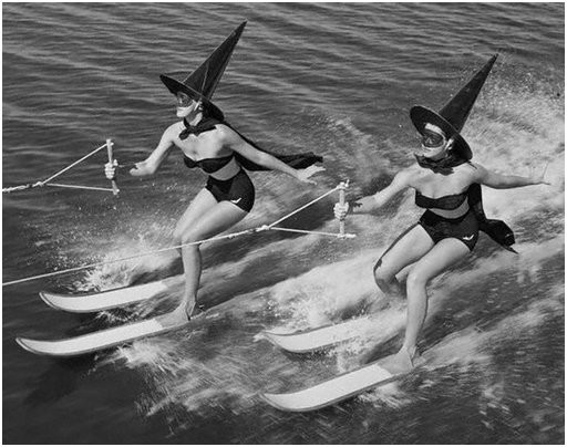 Black and white photo of two water-skiing women wearing black bikini swimsuits, capes, masks, and pointy witch hats.