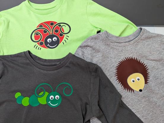 Three children's t-shirts, one bright green with a ladybug (ladybird), one dark grey with a green caterpillar, and one light grey with a hedgehog.