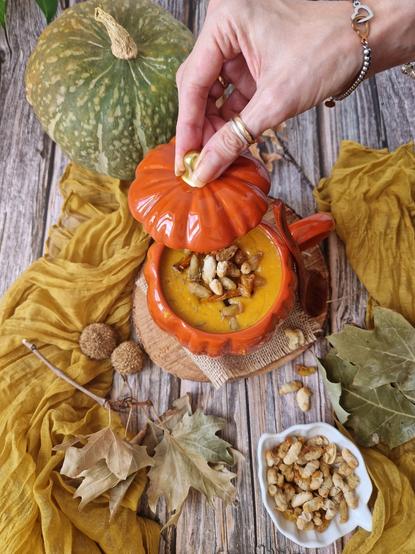 Una Vellutata di zucca con sopra dei cannellini croccanti fatti al forno, all'interno di una ciotola a forma di zucca.
Intorno varie foglie secche, una zucca delica intera e un panno color ocra