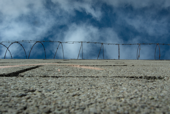 photograph of a wall looking up to see barbwire and beyond blue sky