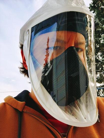 Me, wearing papr with white hood over black n95, and brown hoodie; standing outside in front of blueish does and coniferous trees