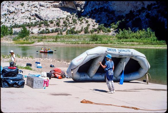 A large grey inflatable raft is propped up by an oar, partially upside-down. It sits on a concrete boat ramp adjacent to a greenish river. Piles of equipment - dry bags, coolers, life preservers - sit to the left. Near the raft, a woman stands facing away from the camera. She is wearing a blue bucket hat, blue-striped shirt, and grey pants. A blue sweatshirt is tied around her waist. In the river beyond, another raft approaches the boat ramp. Beyond the river, rocky cliffs rise steeply.