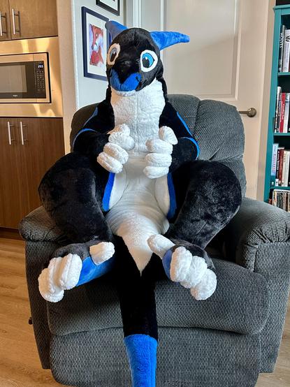 Setting: home interior, kitchen/living area. Photo of a large (80", 200cm) friendly looking plush dragon sitting in a recliner.
